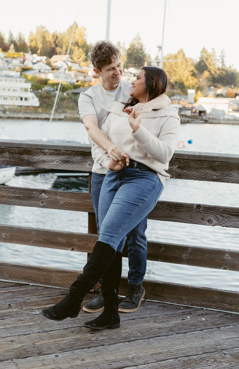 A couple shares a romantic moment together on a wooden dock overlooking a marina with boats and trees in the background.