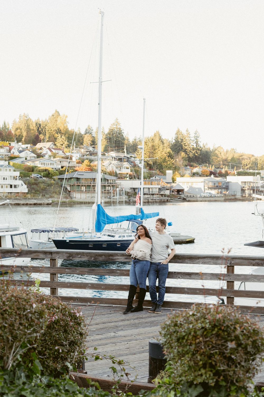 Couple enjoying a romantic moment together on a wooden dock with boats and autumn trees in the marina background.