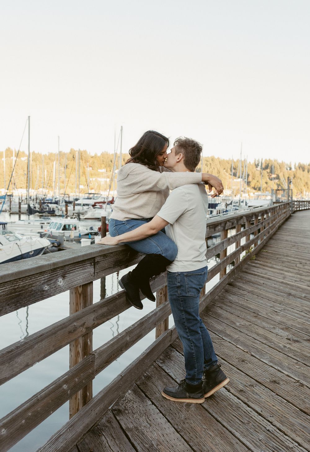 A romantic moment unfolds between two people embracing on a wooden pier overlooking a marina at sunset.