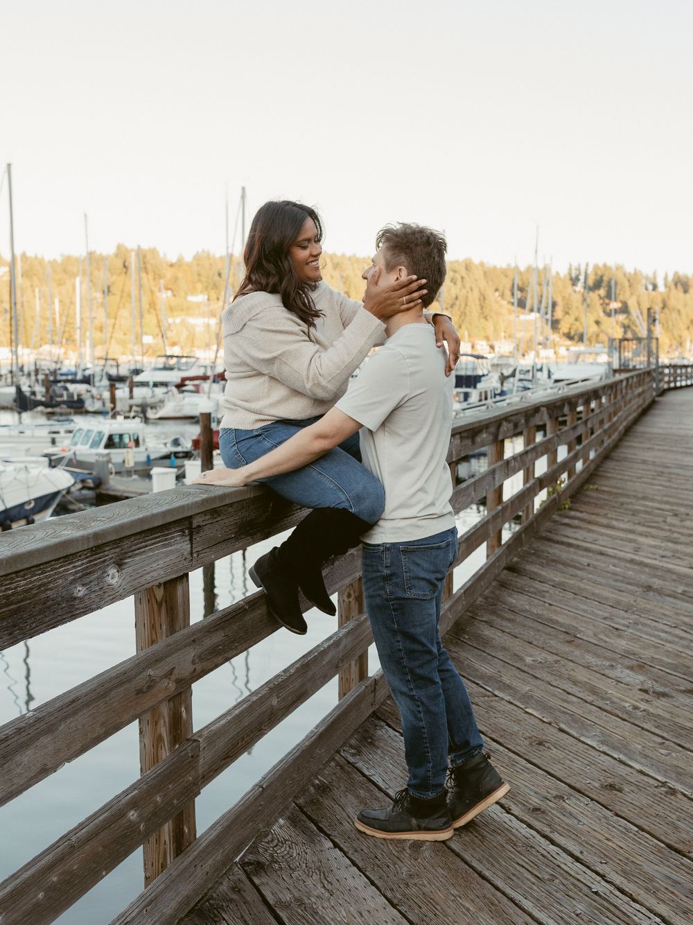 Couple embracing on waterfront boardwalk with sailboats and marina in late afternoon light.