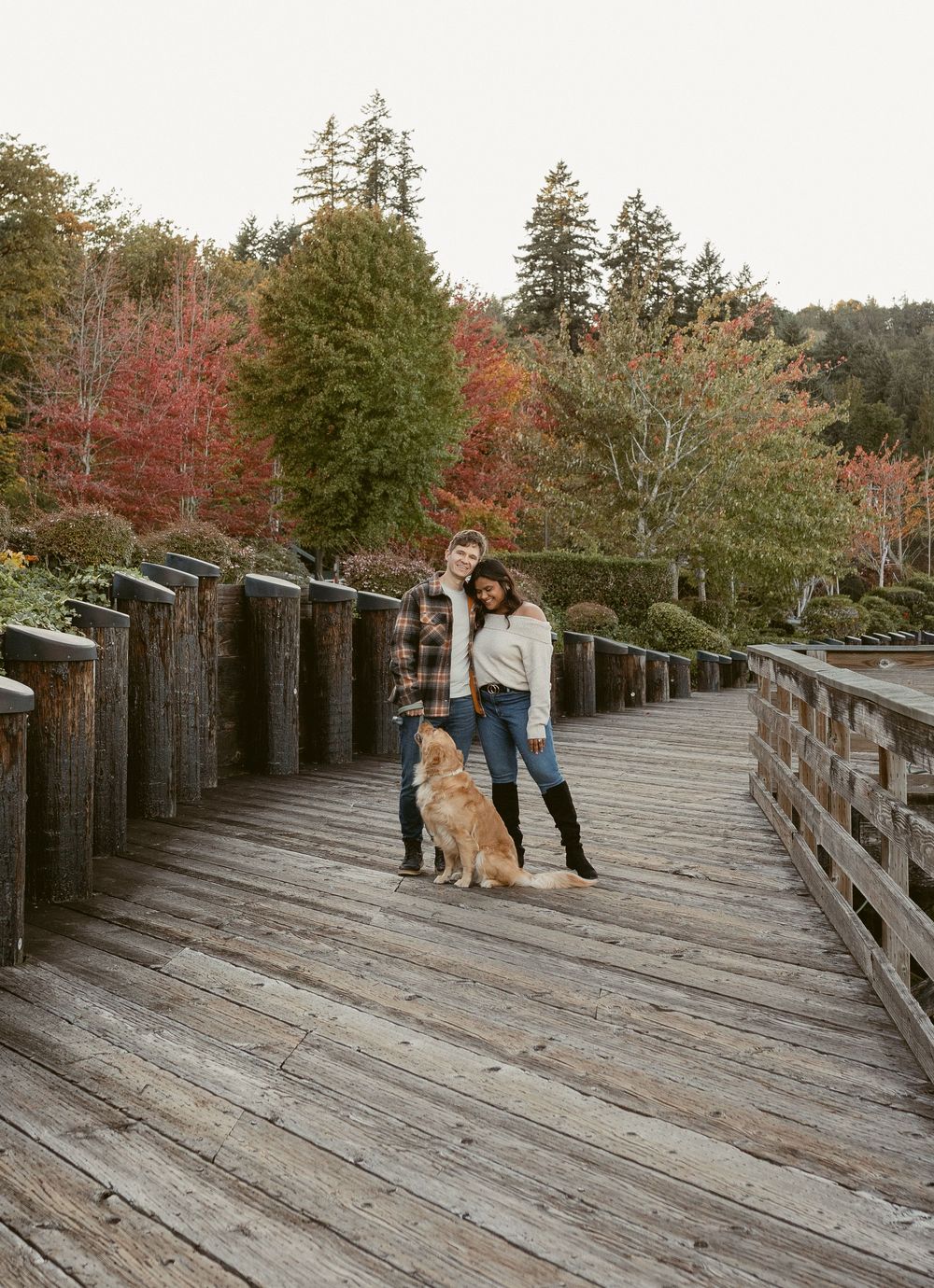 A couple and their golden retriever pose together on a wooden boardwalk surrounded by autumn trees.
