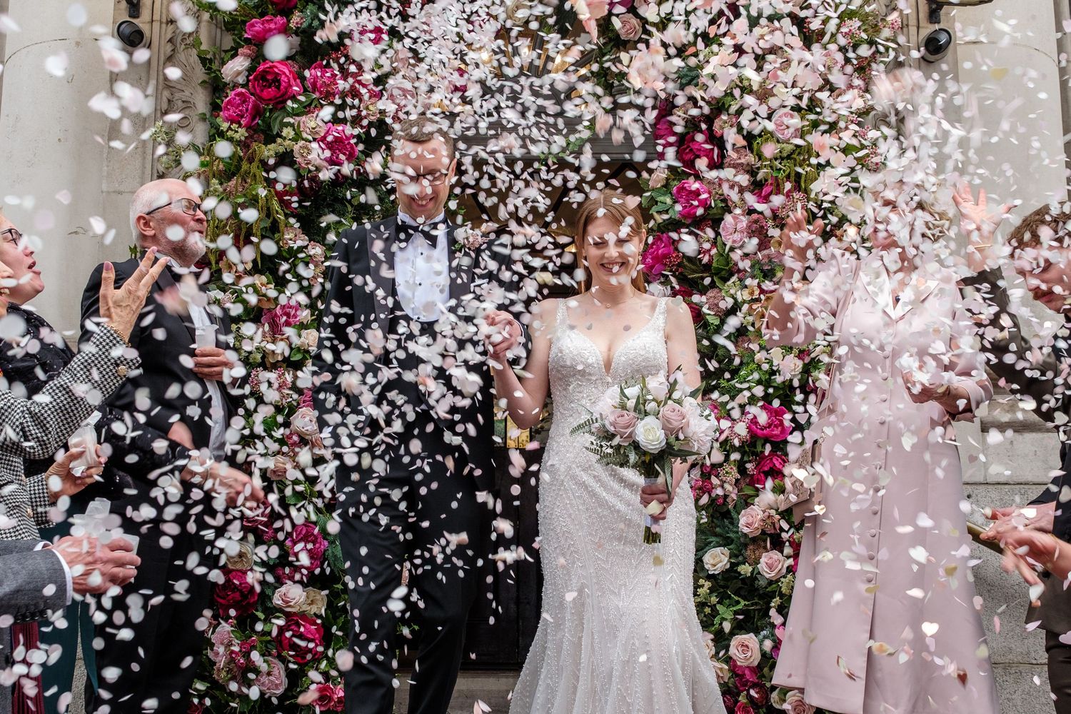 Wedding celebration with guests throwing white flower petals and confetti during outdoor ceremony under floral arch.