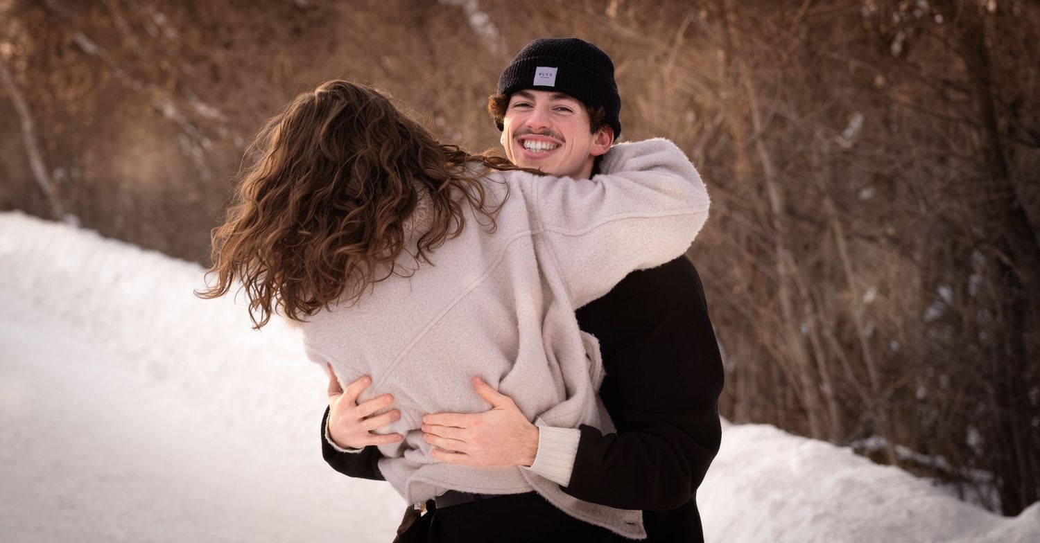 Romantic winter scene with couple embracing in falling snow among trees.
