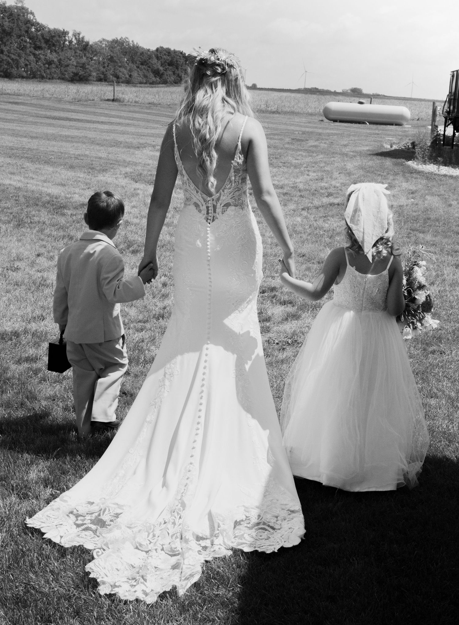 Black and white photo showing a bride in a fitted lace gown walking with two children in formal attire on a grassy field.