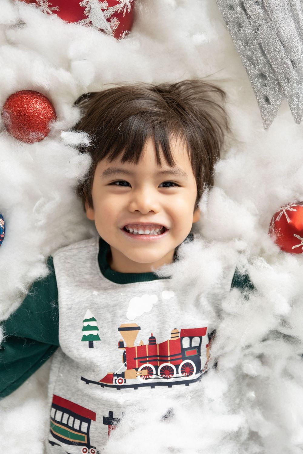 Child wearing holiday sweater with train design lies on white fluffy background with red ornaments.