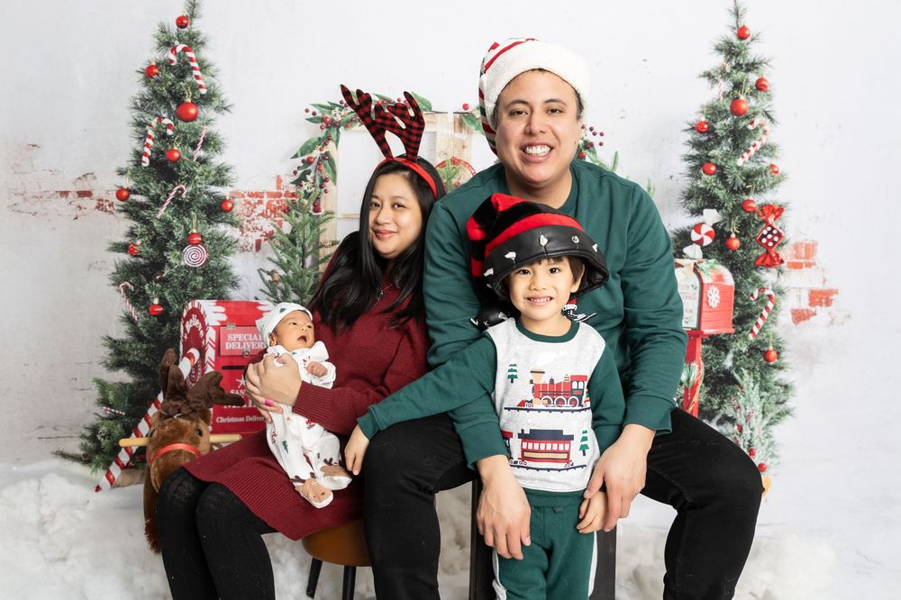 Family wearing matching Christmas pajamas poses together in front of decorated trees in white studio setting.