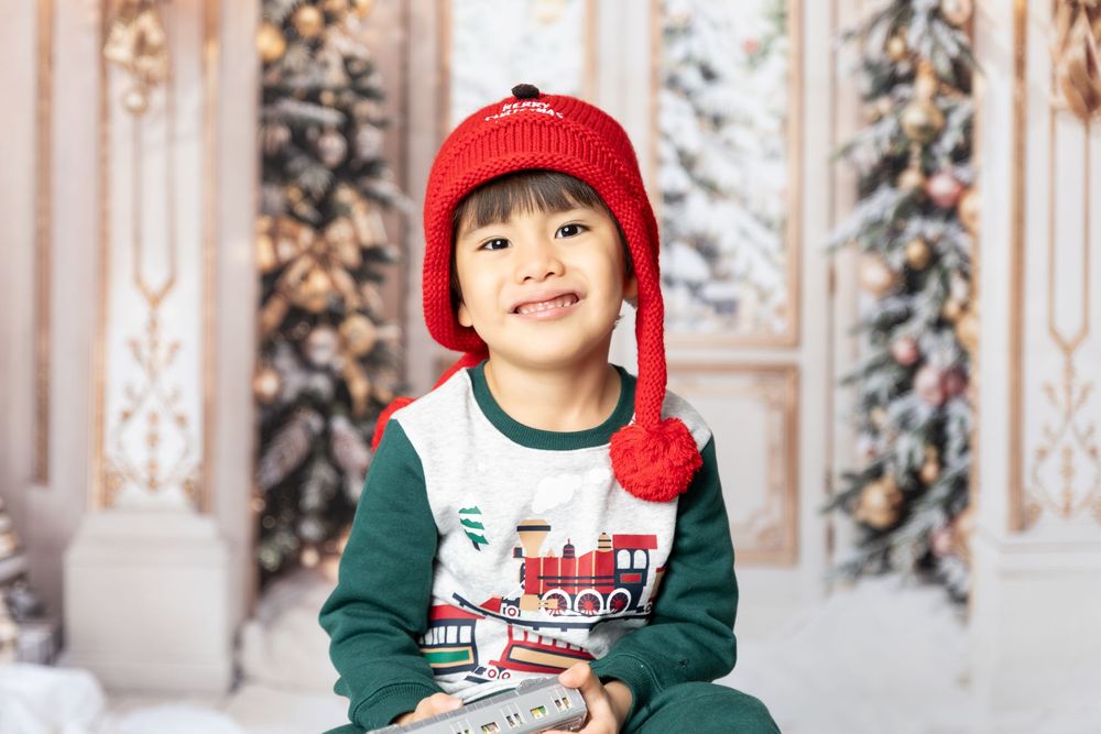 A toddler in a red winter hat and green sweater smiles while sitting in a festive holiday setting with snowy trees.