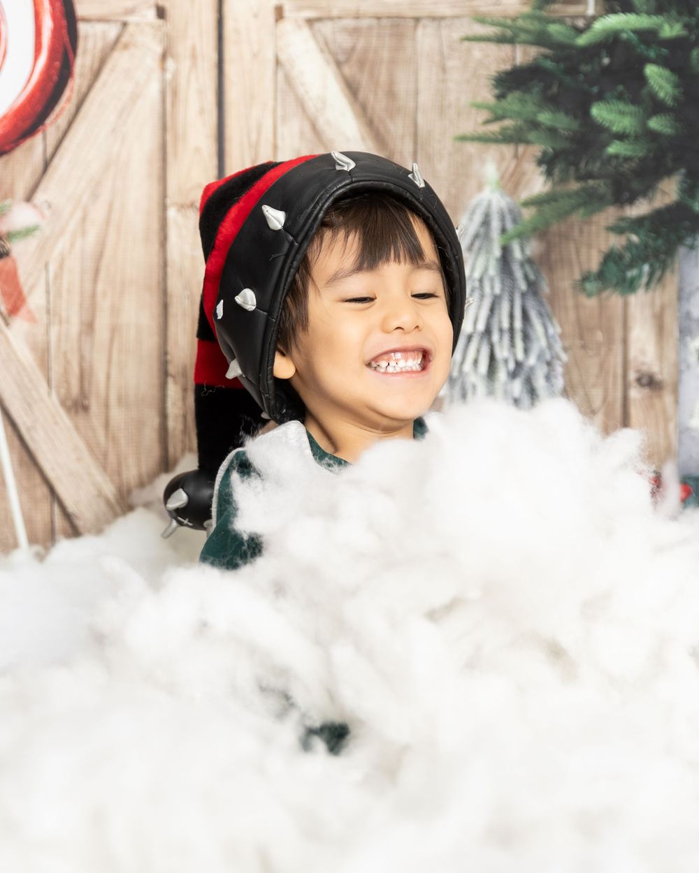 A young child in a red hat smiles joyfully while playing in a snowy winter scene with Christmas decorations.