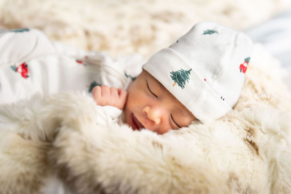 A newborn baby wearing a white knit hat sleeps peacefully on soft cream-colored bedding.