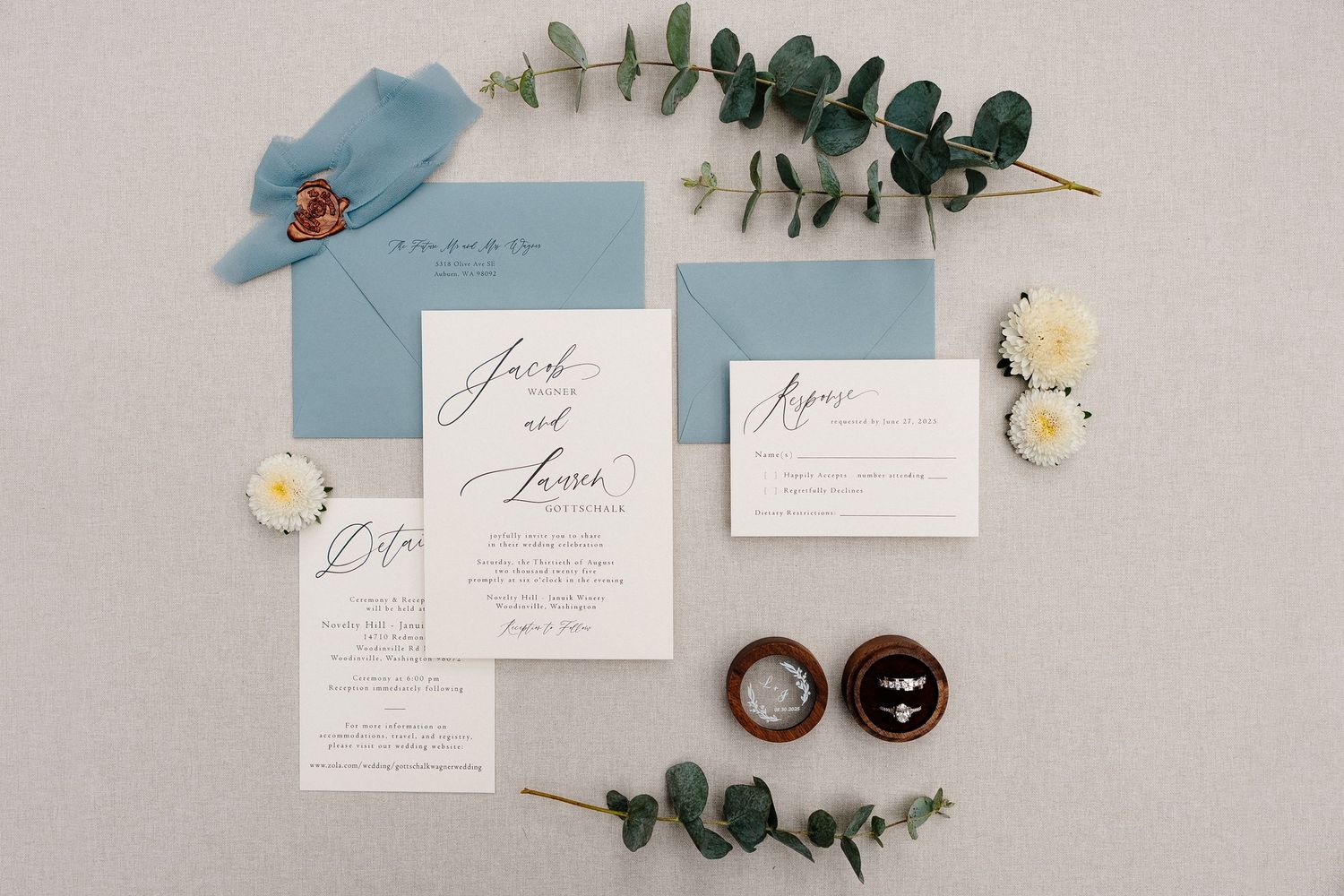 Elegant wedding invitation suite with white and blue stationery, eucalyptus sprigs, and small white roses on gray background.