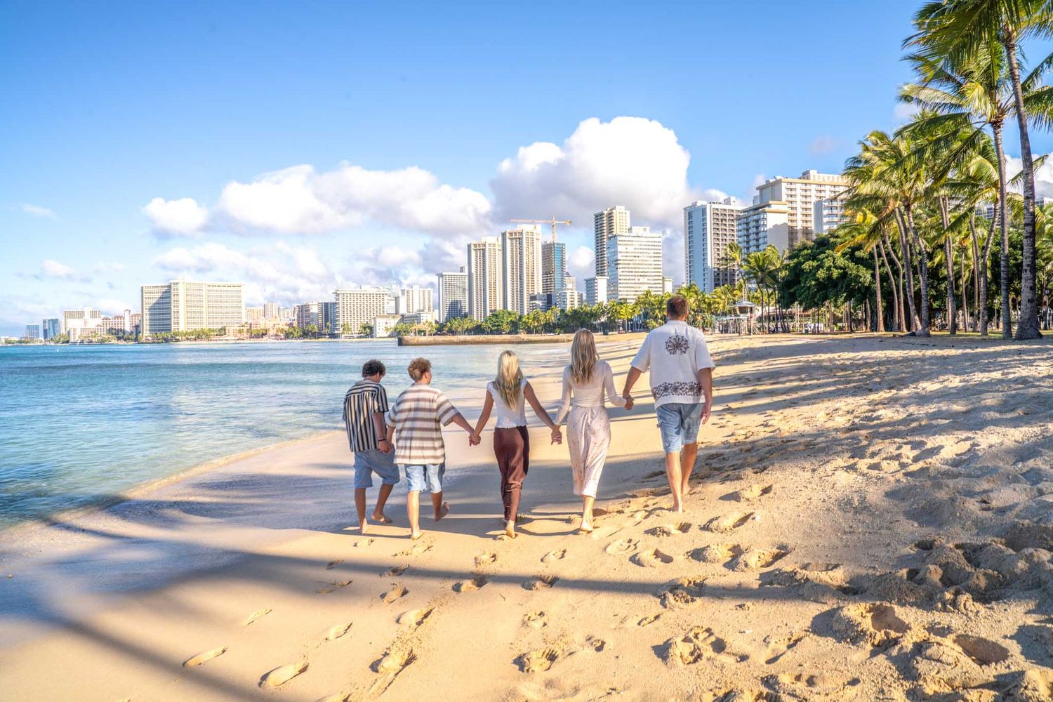 Family photo session at Queen’s Beach, framed by palm-lined paths, sandy Waikiki shores, blue ocean, and iconic high-rise backdrops.