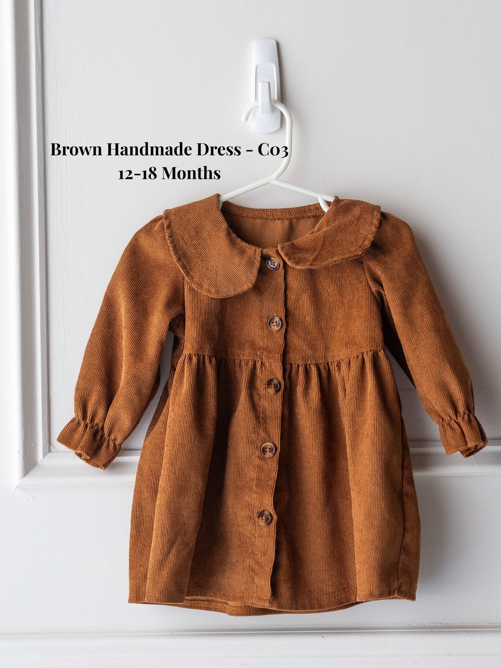Brown corduroy baby dress with peter pan collar and button front design for 12-18 months size hanging on white hanger.