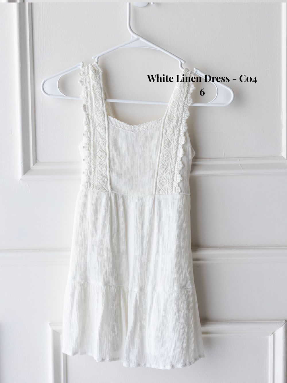 White sleeveless cotton linen dress with delicate embroidery details hanging on white hangers against white wall.