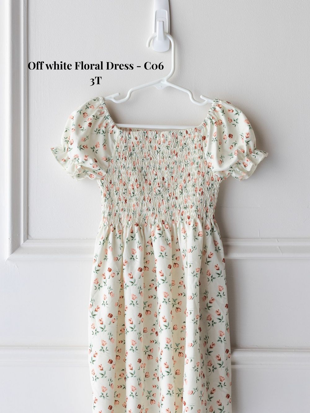 White floral smocked dress with puff sleeves and delicate pink rose print hanging on white wall display.
