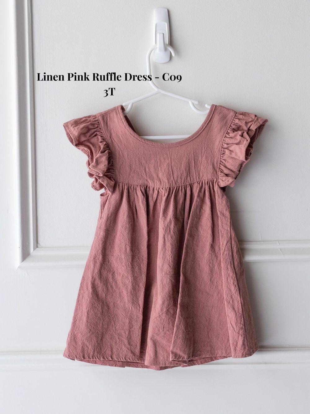 Pink linen ruffle dress with flutter sleeves hung against white wall.