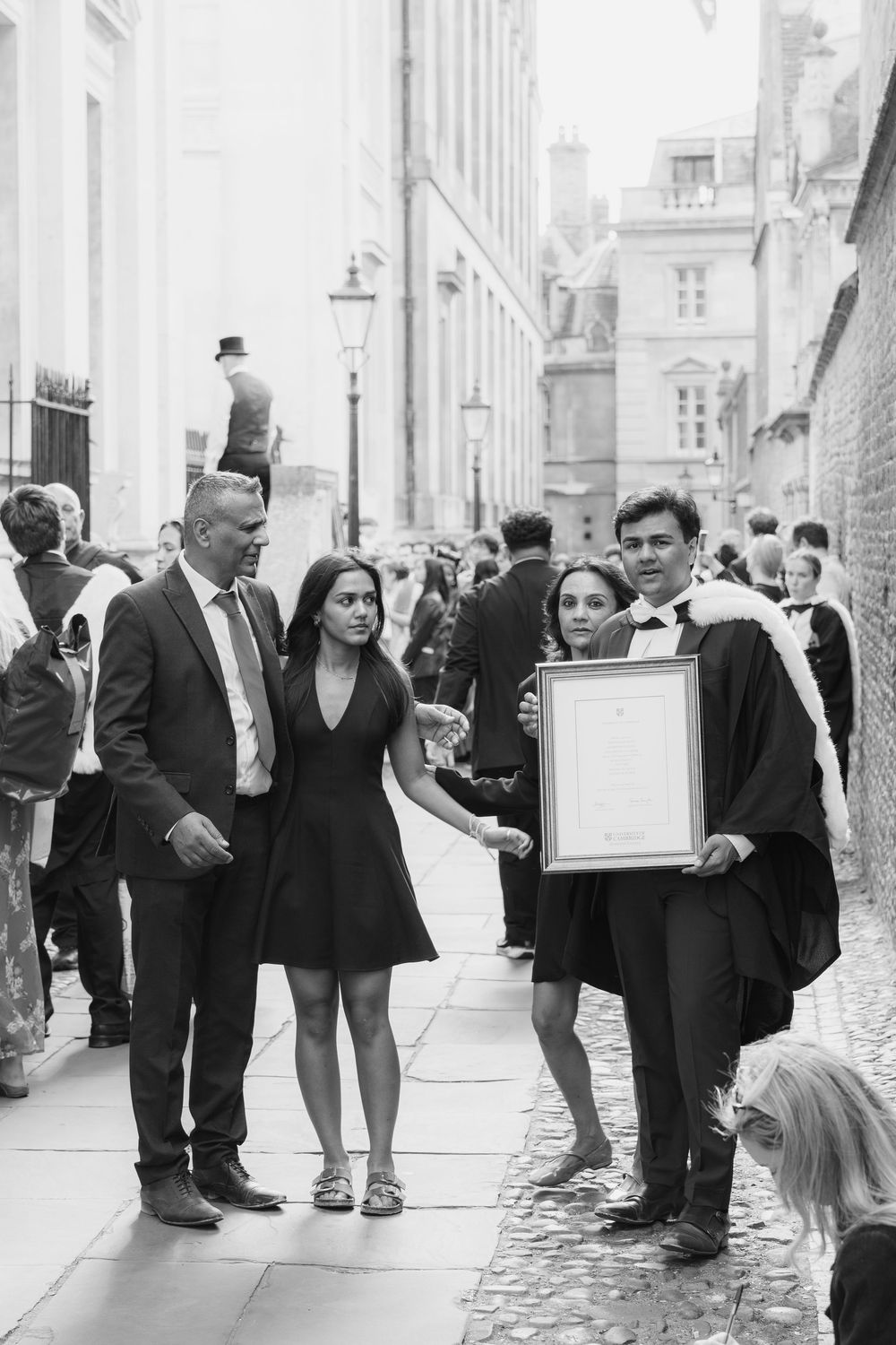 People in formal attire gather outside a historic building with stone architecture and cobblestone streets.