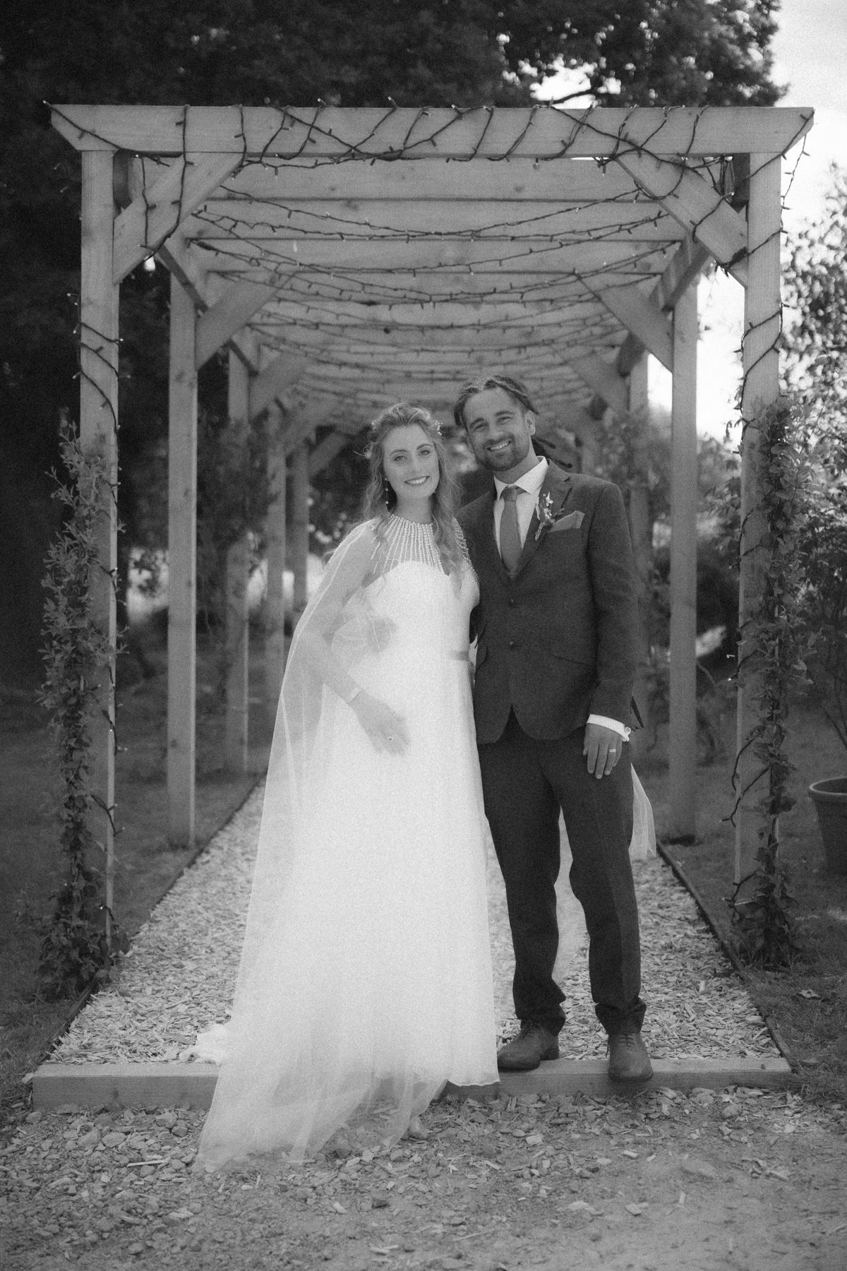 A black and white wedding photo showing a couple under a garden arbor decorated with vines and branches.