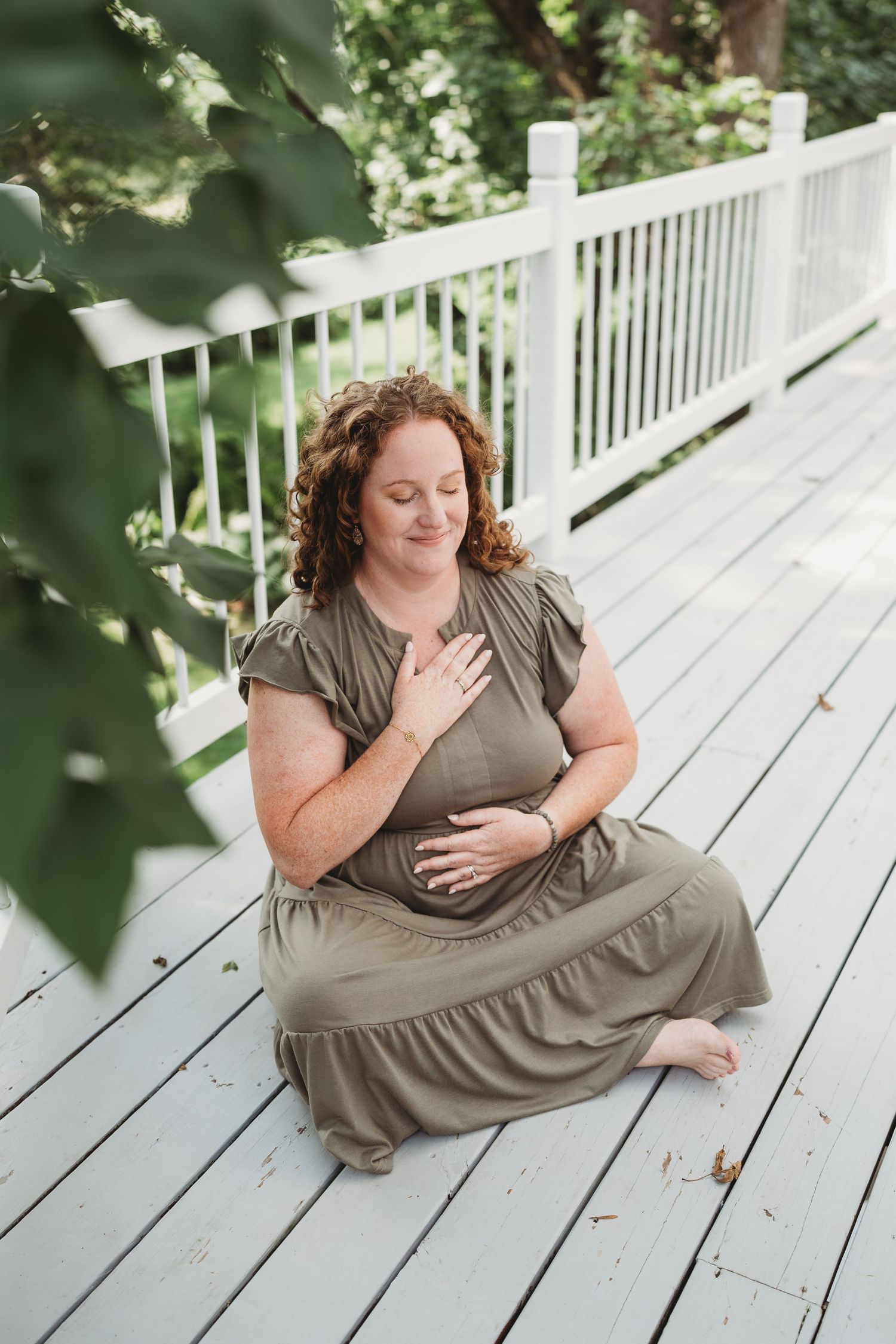 A person in an olive dress sits on a white wooden deck surrounded by greenery and natural light.