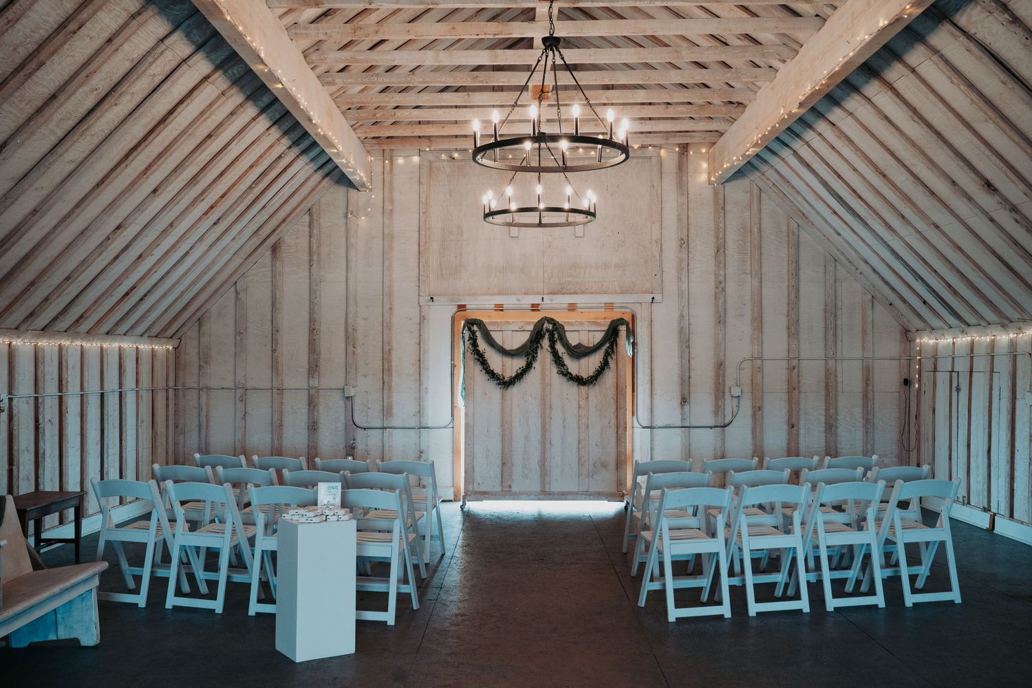 Rustic barn wedding venue interior with white walls, exposed beams, and rows of blue chairs set up for ceremony.