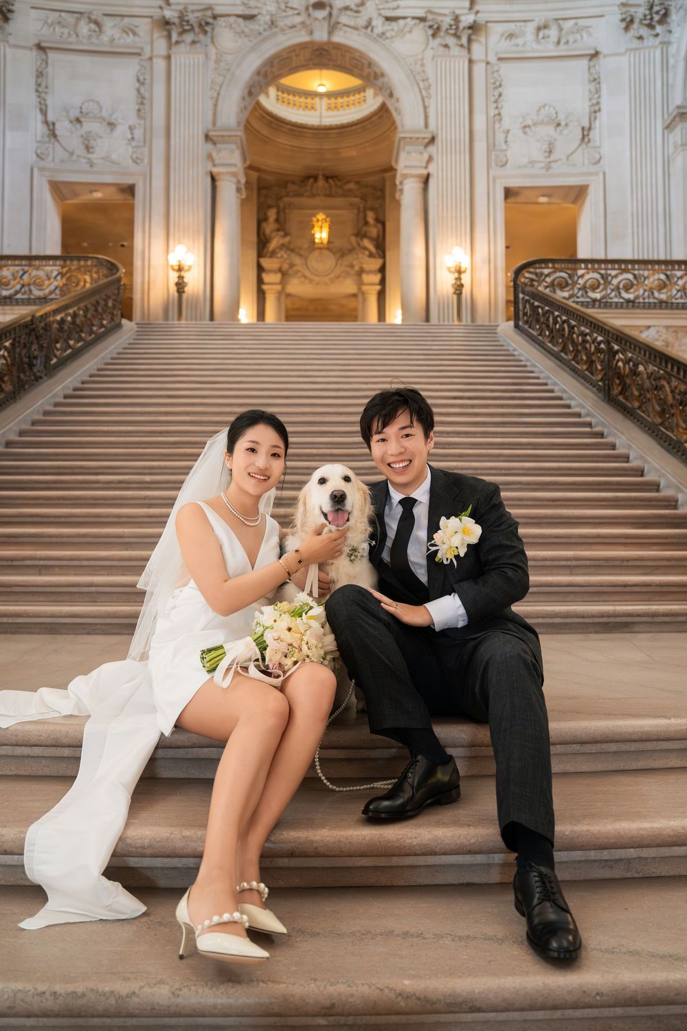 Newlyweds sit playfully on grand marble steps in an ornate building with decorative railings.