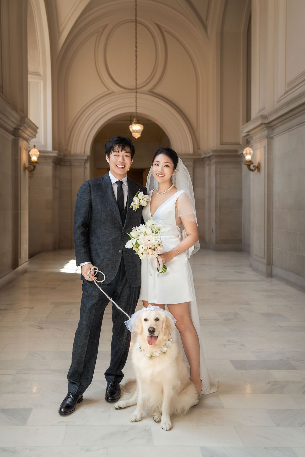 Wedding couple poses with their golden retriever in an elegant marble hallway with arched ceilings and warm lighting.