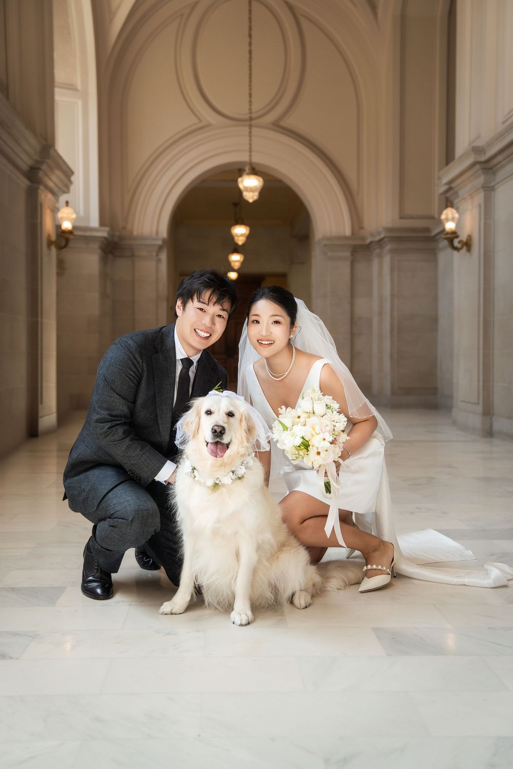 Wedding couple poses with their white dog in an elegant hallway with arched ceilings and columns.