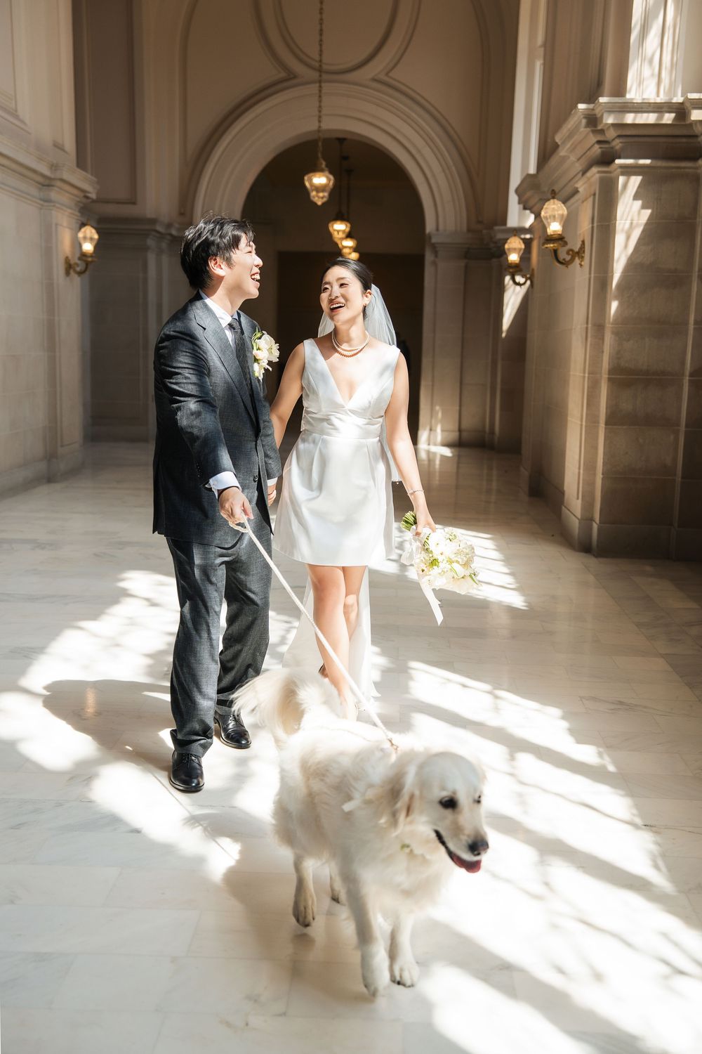 Wedding couple walks with their white dog in a sunlit hallway with beautiful architectural details.