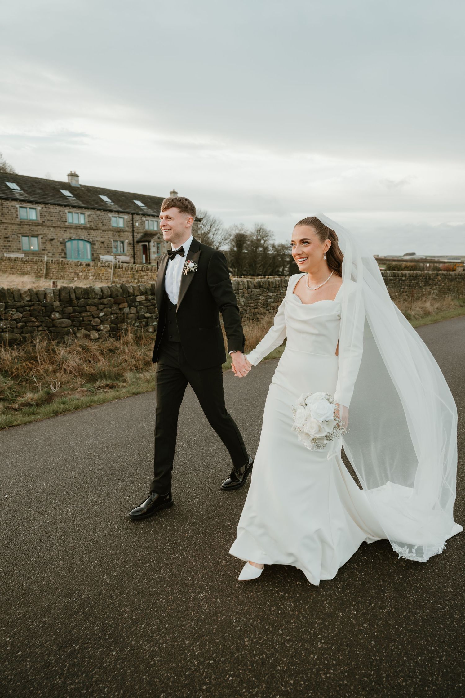 A couple walks together on a country road while the bride's white dress and veil flow in the wind.