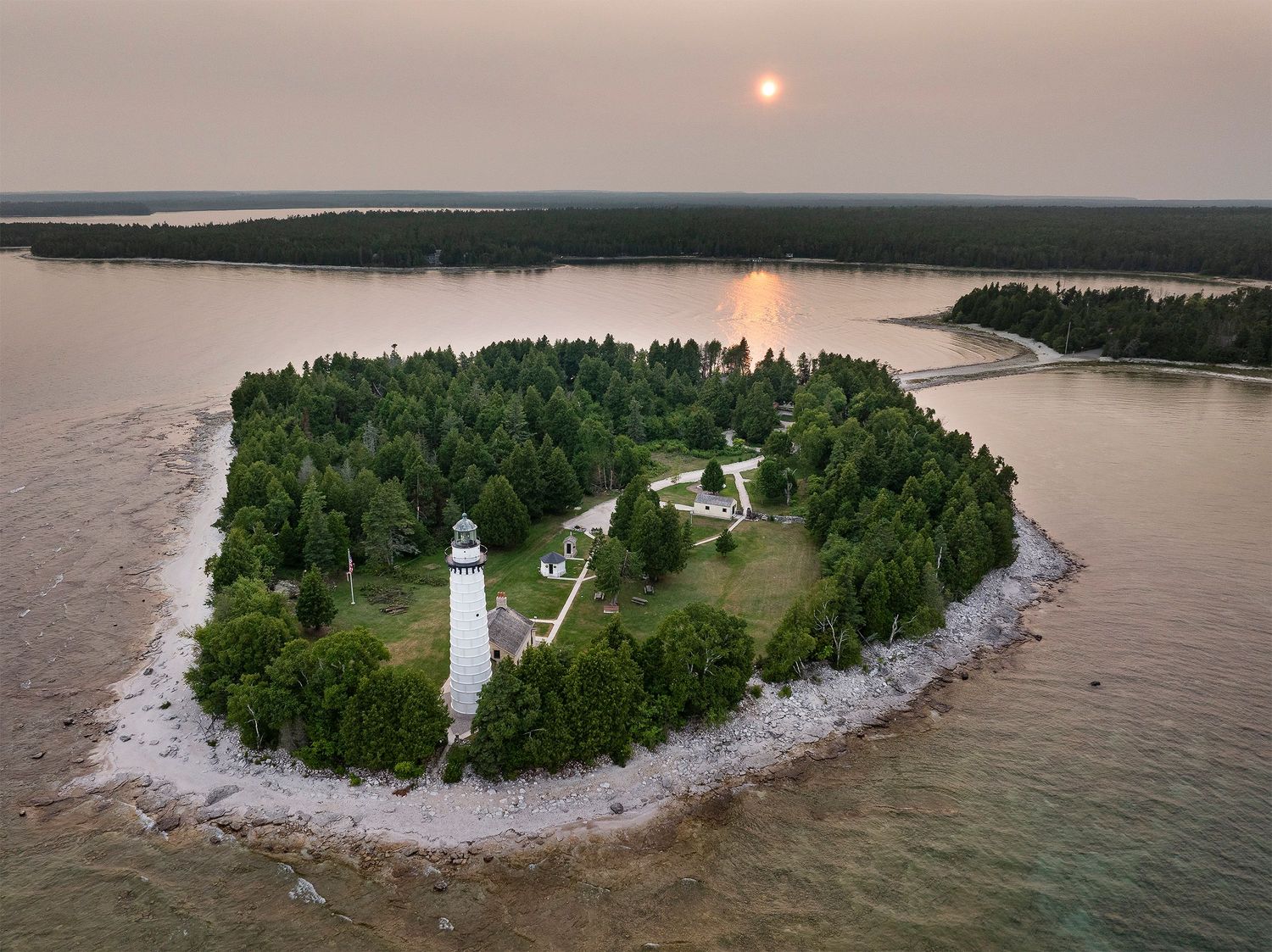 Aerial view of a lighthouse on a small forested island surrounded by turquoise waters during sunset.