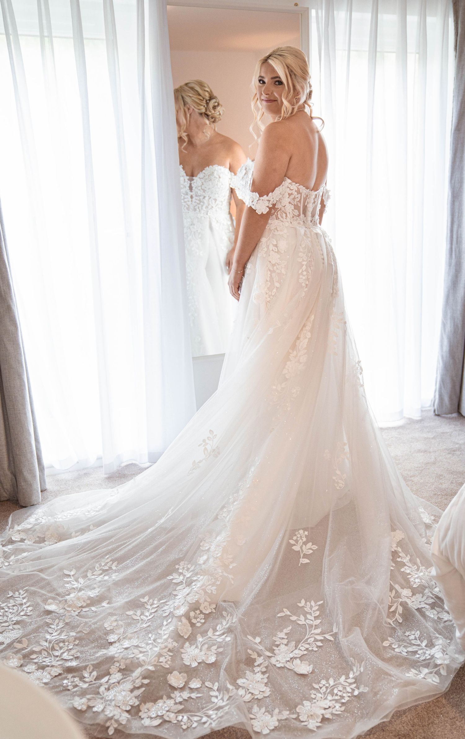 Elegant off-shoulder wedding gown with delicate lace appliques and flowing train captured in front of sheer curtains.