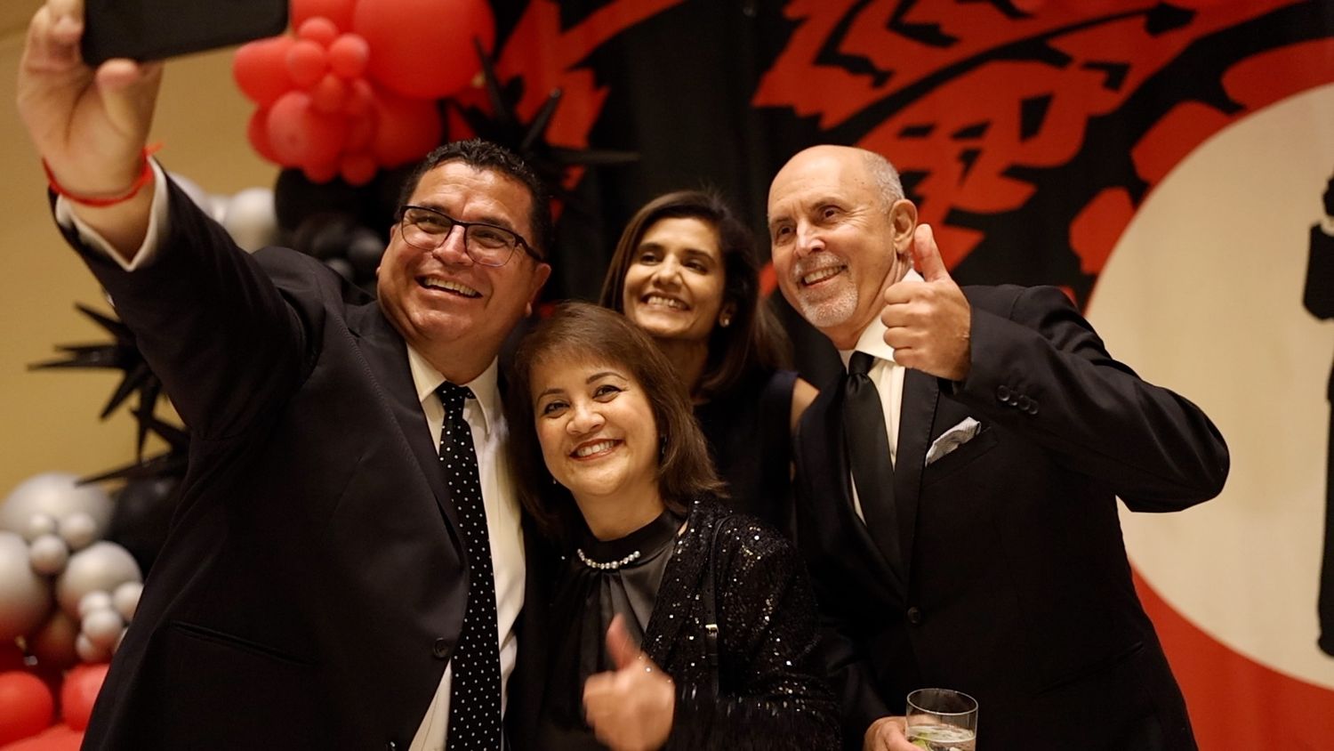 A group celebrates together at a festive event with red decorations and balloons in the background.