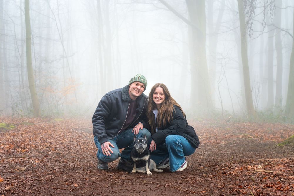 Couple with dog posing in misty autumn forest on a leaf-covered path, surrounded by fog-filled trees.