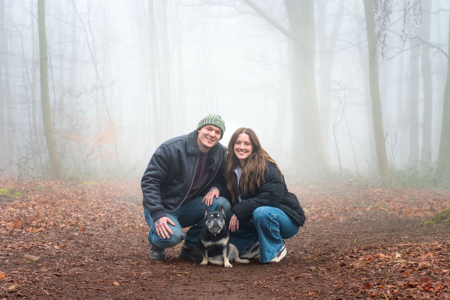 Couple with dog posing in misty autumn forest on a leaf-covered path, surrounded by fog-filled trees.