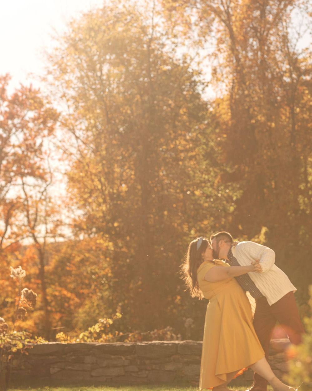 Couple embraces during sunset in autumn setting with golden light filtering through trees.