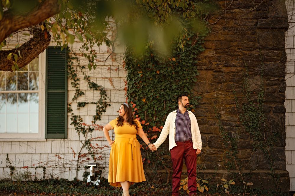 Couple in yellow dress and burgundy pants pose against ivy-covered stone wall.