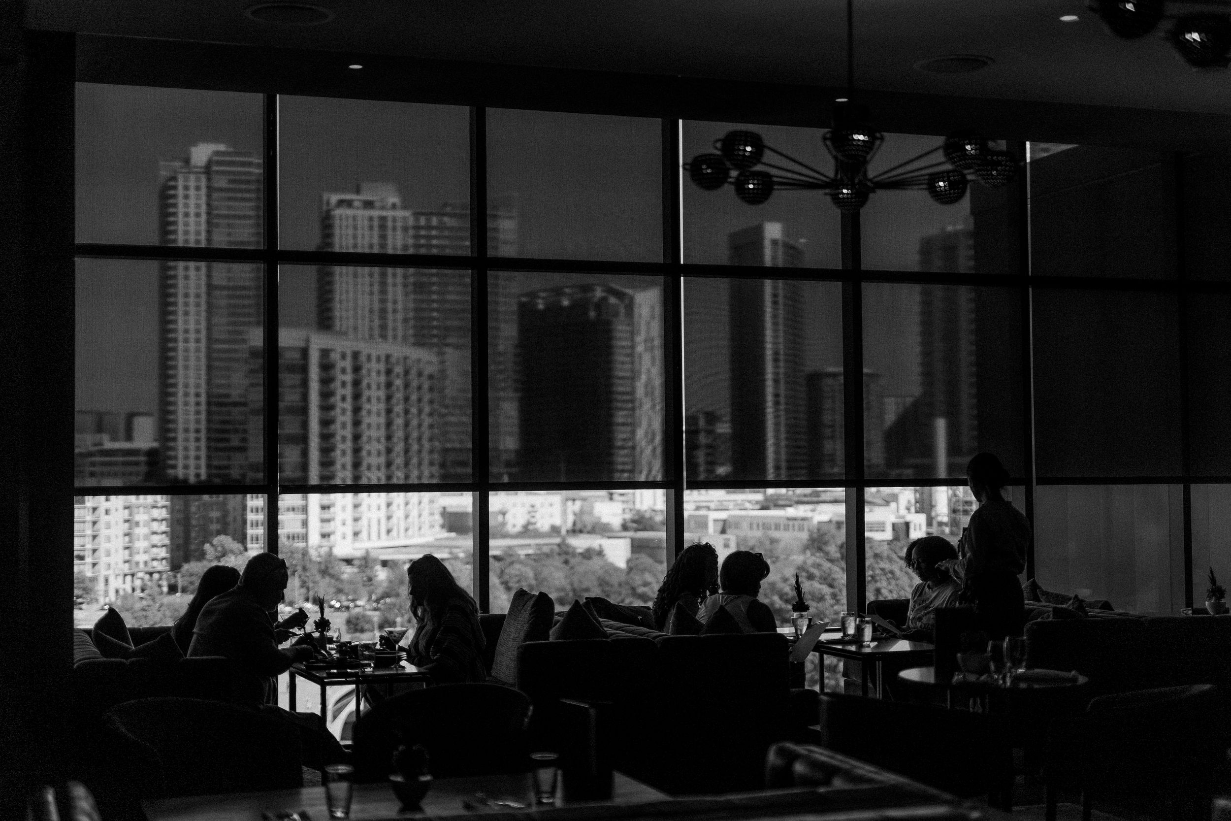 Silhouettes of diners in a dark restaurant with city skyline visible through large windows in black and white.
