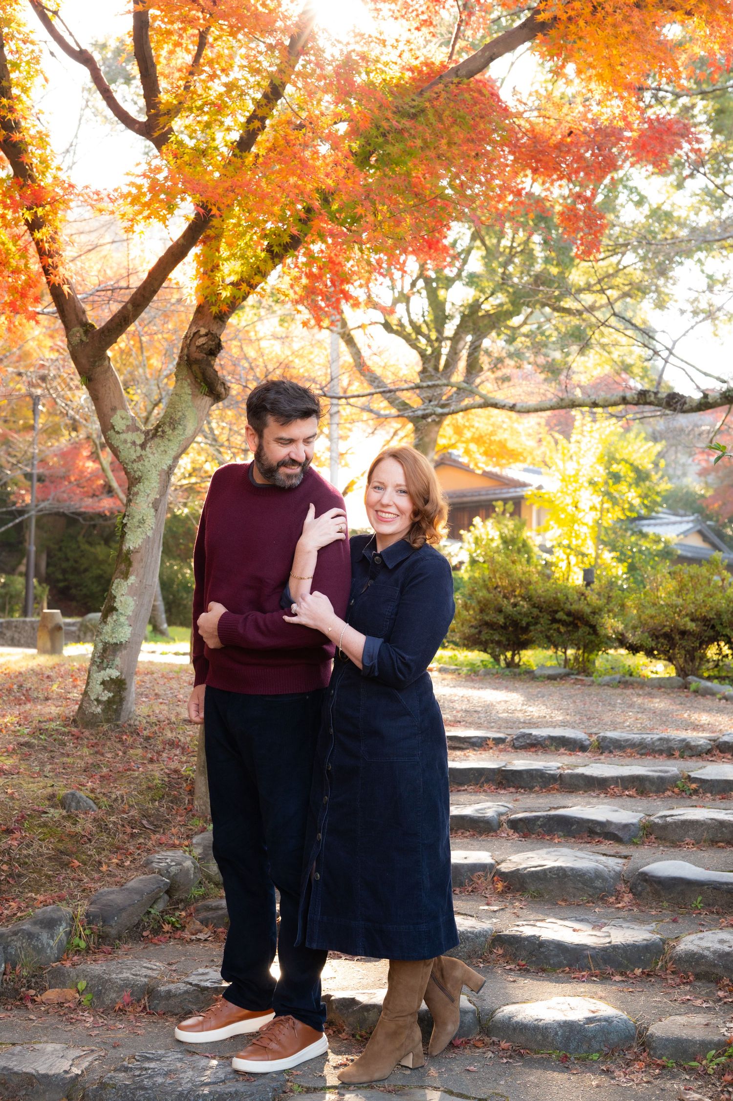 A couple stands together smiling on stone steps beneath vivid orange autumn maple trees.