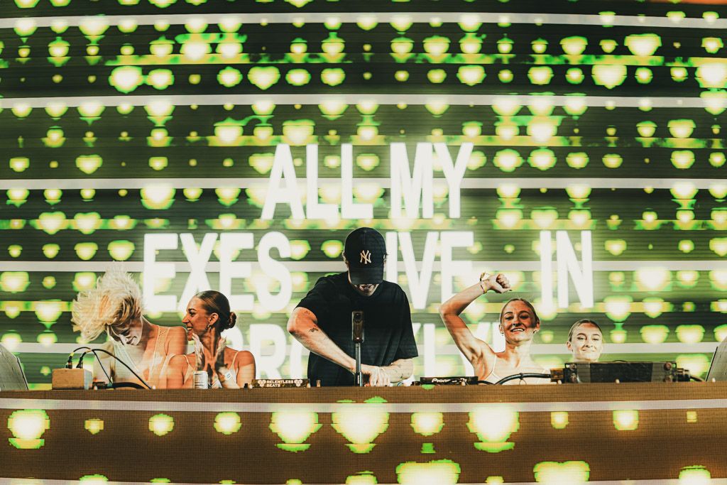 DJs perform at an EDM concert with bright green LED lights and illuminated signage creating an energetic atmosphere.