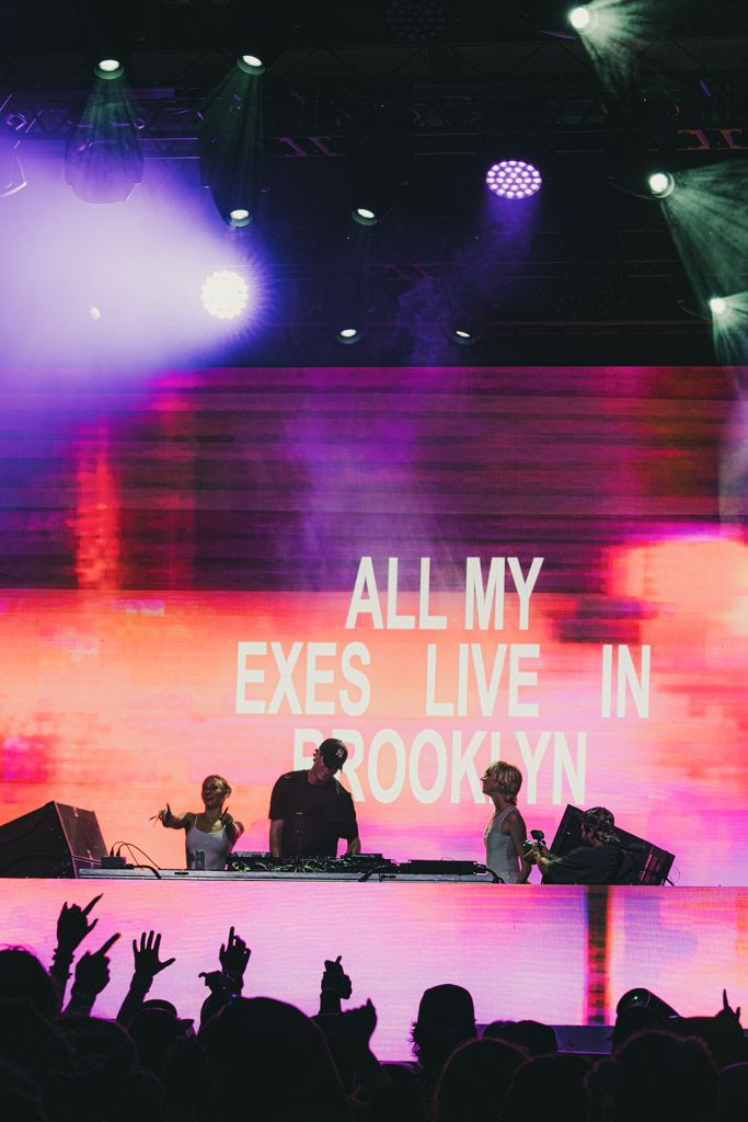 Concert stage with bright pink and purple lighting displaying text on large LED screens as crowd raises hands.