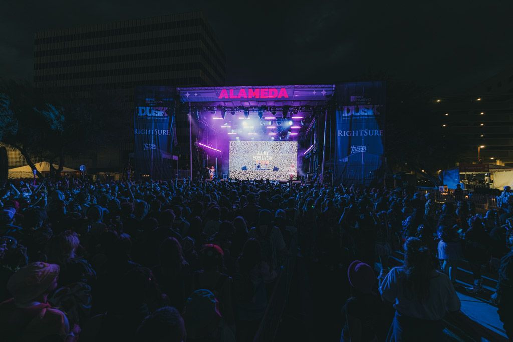 Panoramic shot of a music festival with colorful stage lighting and silhouetted audience members in darkness.