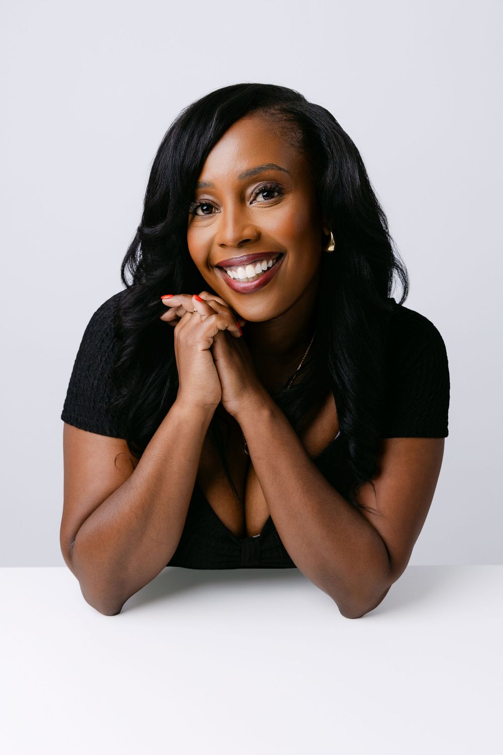Woman in black top poses playfully with hands clasped under chin during studio branding photoshoot.