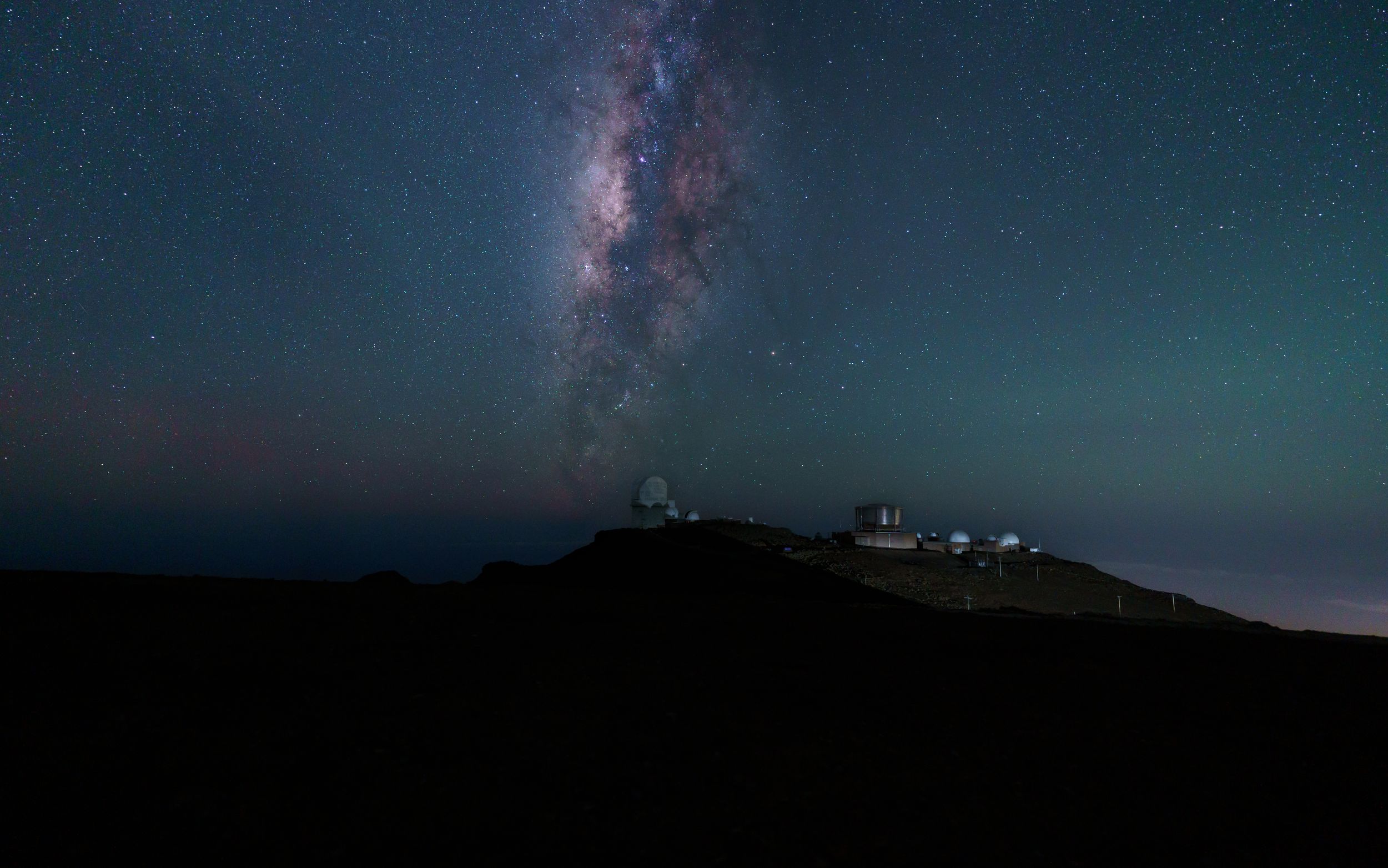 The Milky Way galaxy stretches across the night sky above a silhouetted mountain observatory.