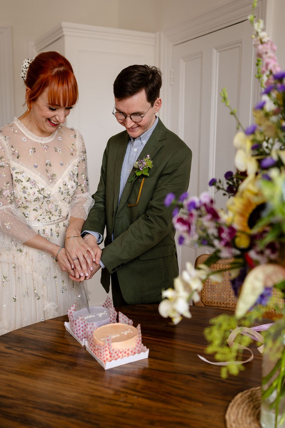 A couple in wedding attire cuts into a pink cake together while colorful floral arrangements decorate the wooden table.
