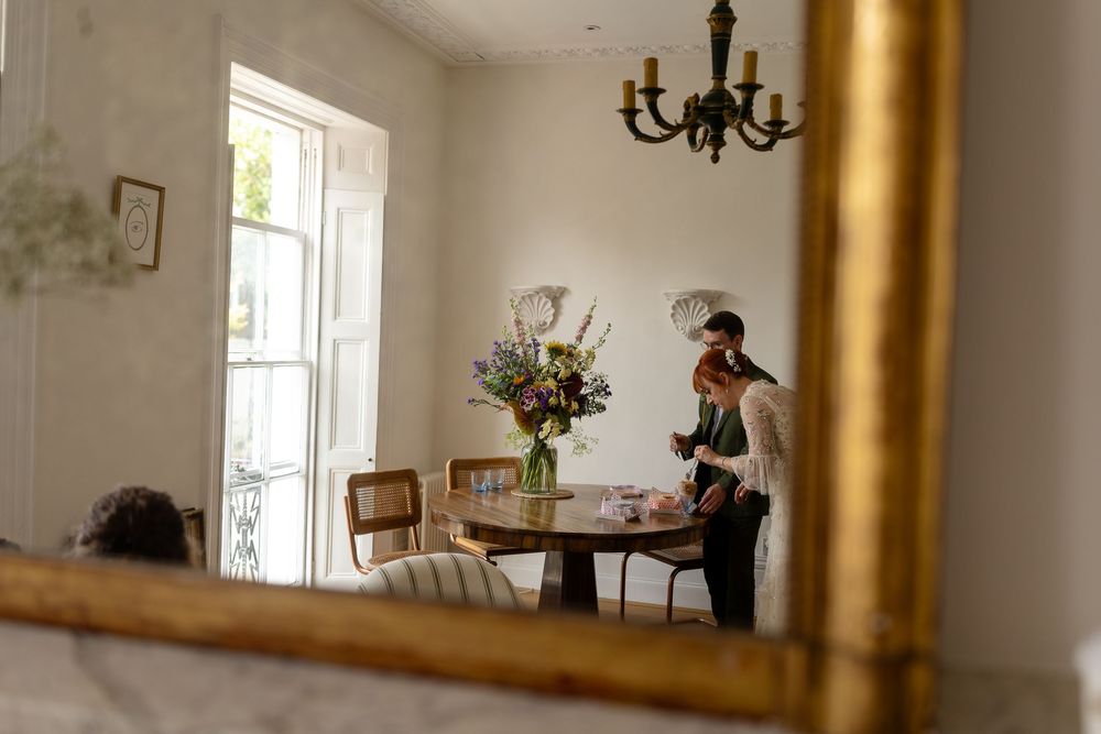 A reflection in a gold-framed mirror shows an elegant dining room with a chandelier and floral arrangement on a table.