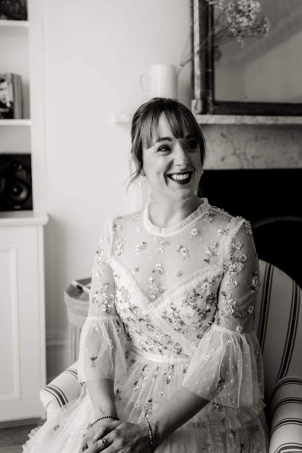 A bride in a sparkly beaded dress with bell sleeves sits in a chair and smiles brightly.