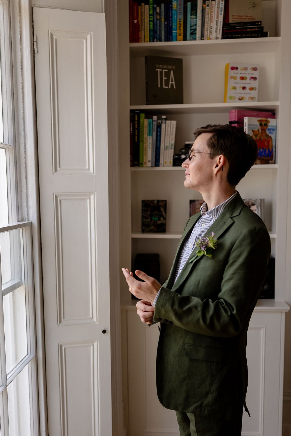 Person in green jacket looking at phone while standing in room with white built-in bookshelves and tea storage.