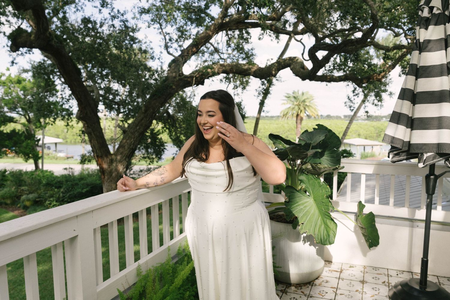 A person in a white dress stands under a tree canopy on a porch during a sunny day.