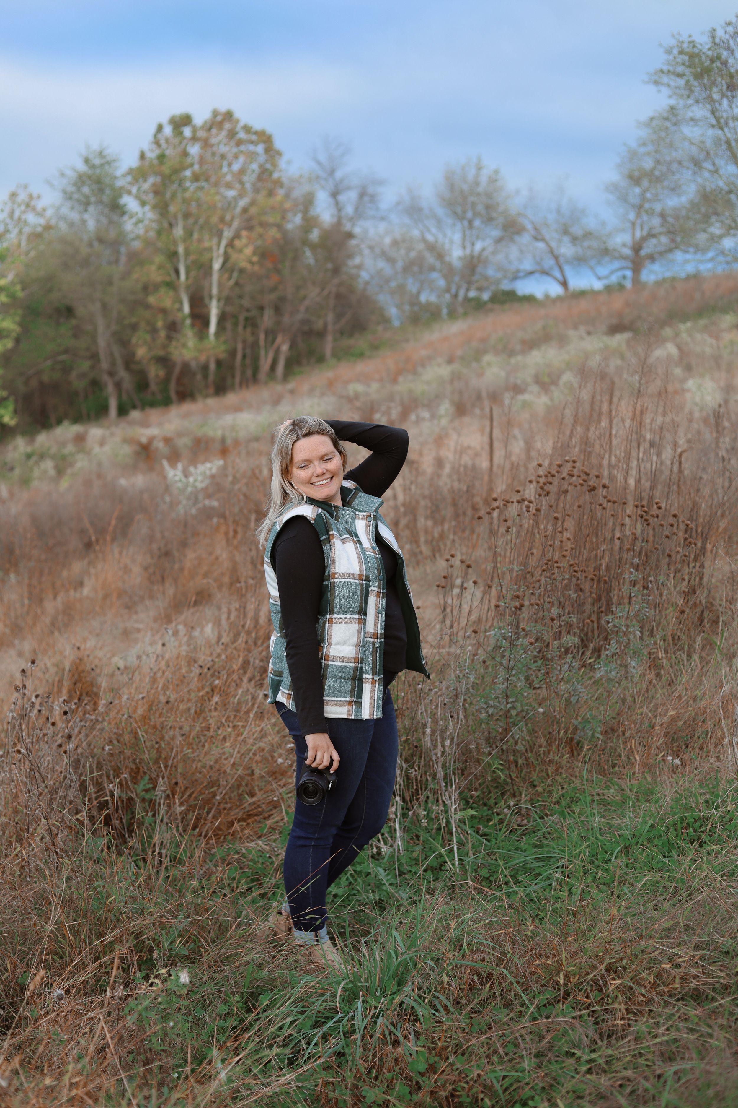 A person in a plaid vest and black sweater poses casually in tall grass during an outdoor photo session.