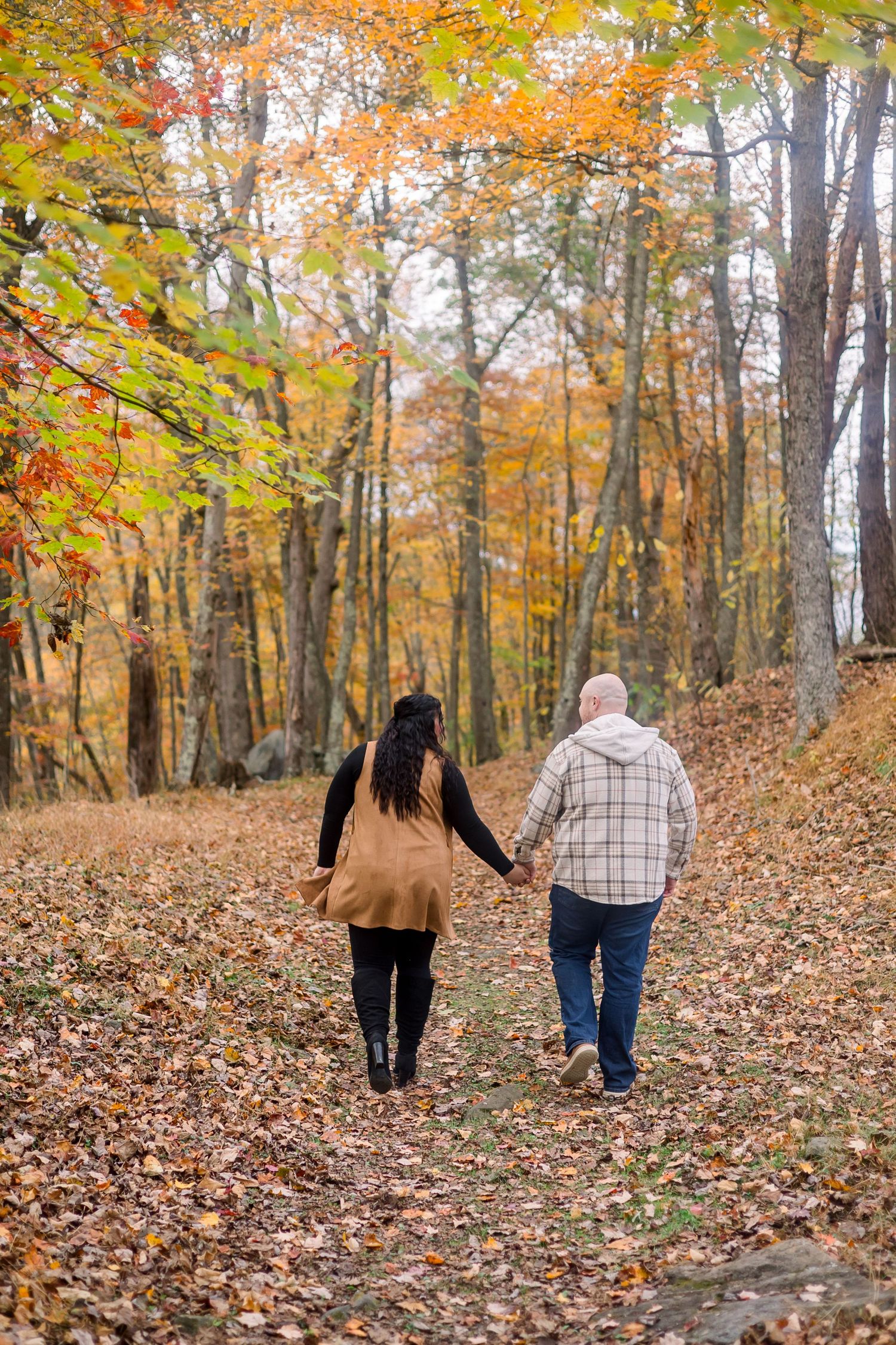 A couple walks hand in hand through a scenic autumn forest path covered in fallen orange and yellow leaves.