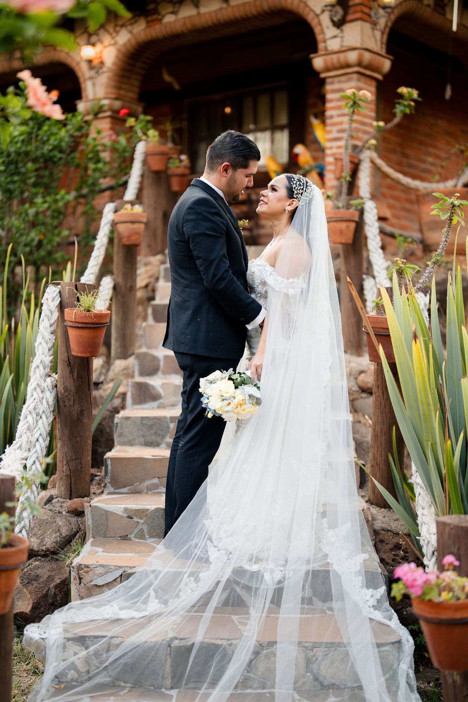 Wedding couple poses in garden setting with flowing veil and rustic architecture.